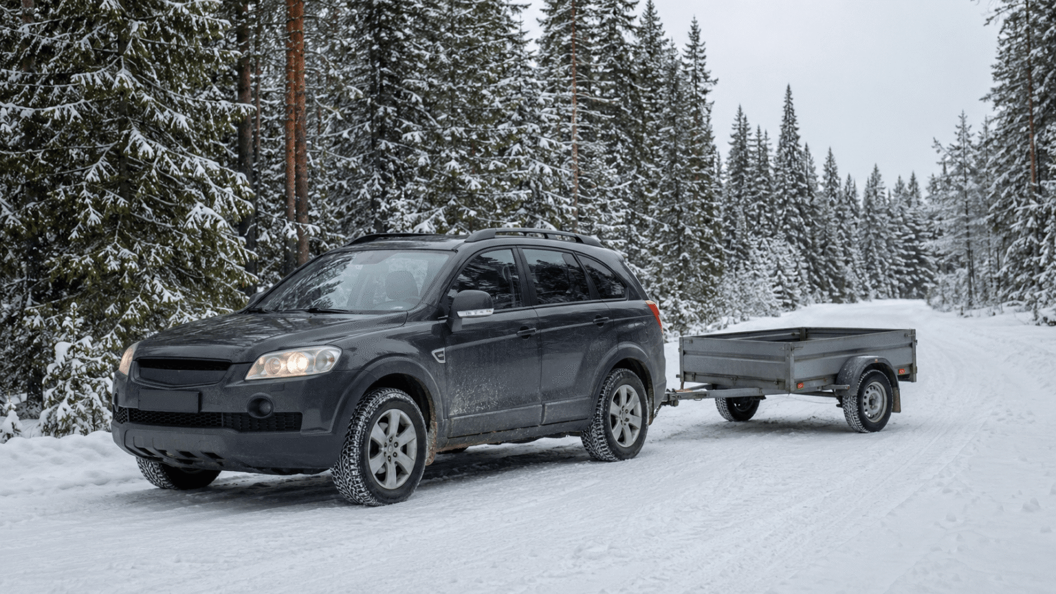 SUV with a small utility trailer attached parked on a snow-covered road in a pine forest.