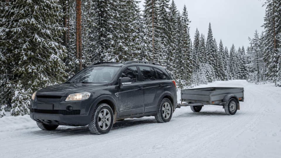 SUV with a small utility trailer attached parked on a snow-covered road in a pine forest.